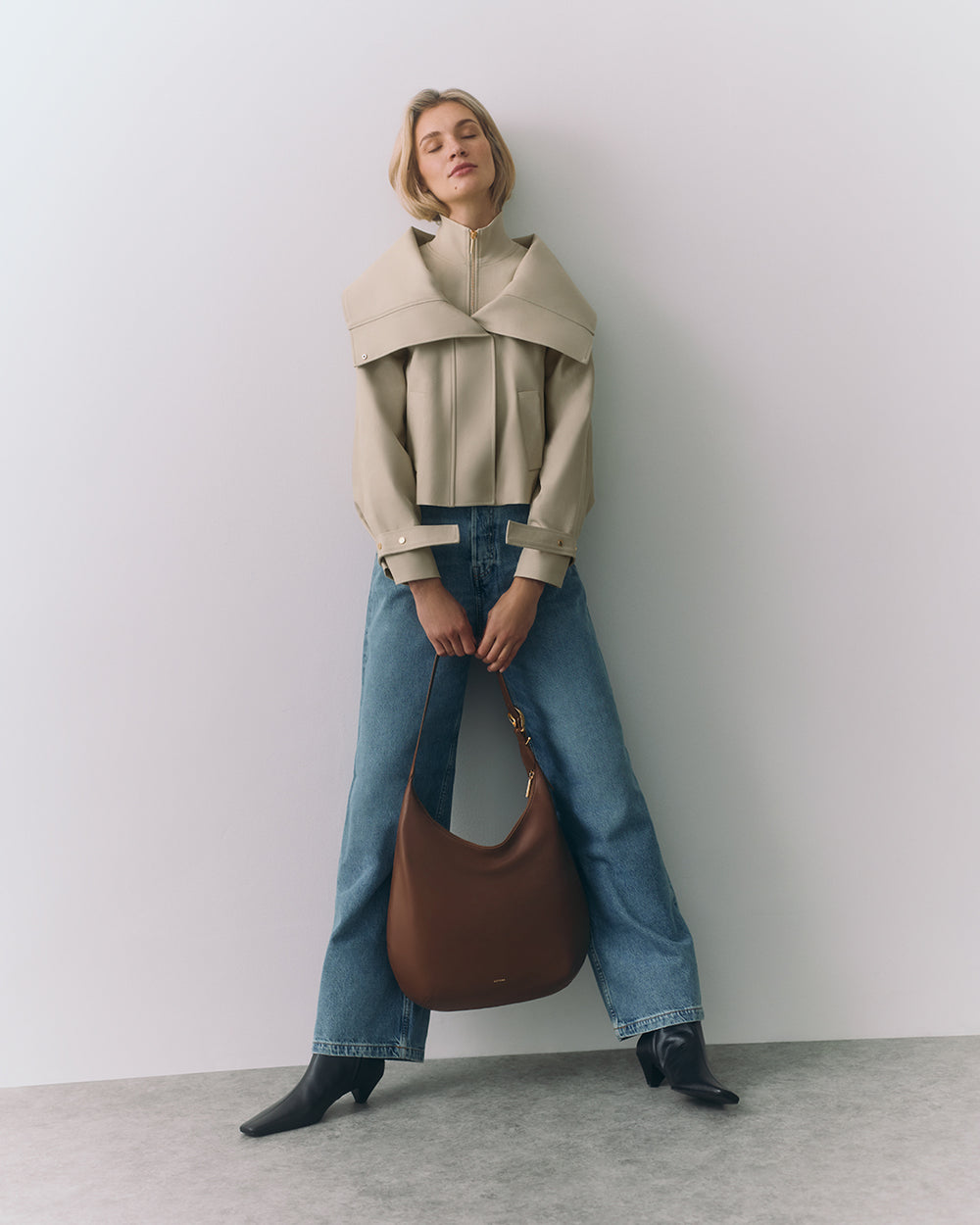 Person in cape and jeans holding a large bag, standing against a plain background.