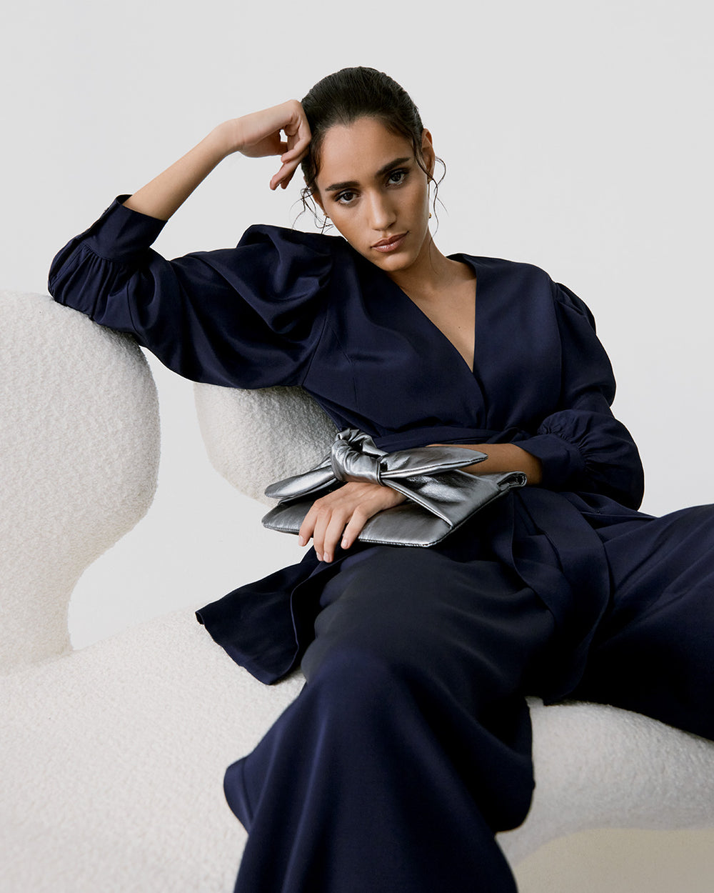 Woman sitting on chair in elegant outfit with hand on head.