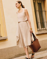 A person in a loose sleeveless top, and flowy skirt holds a large handbag walking by a building.
