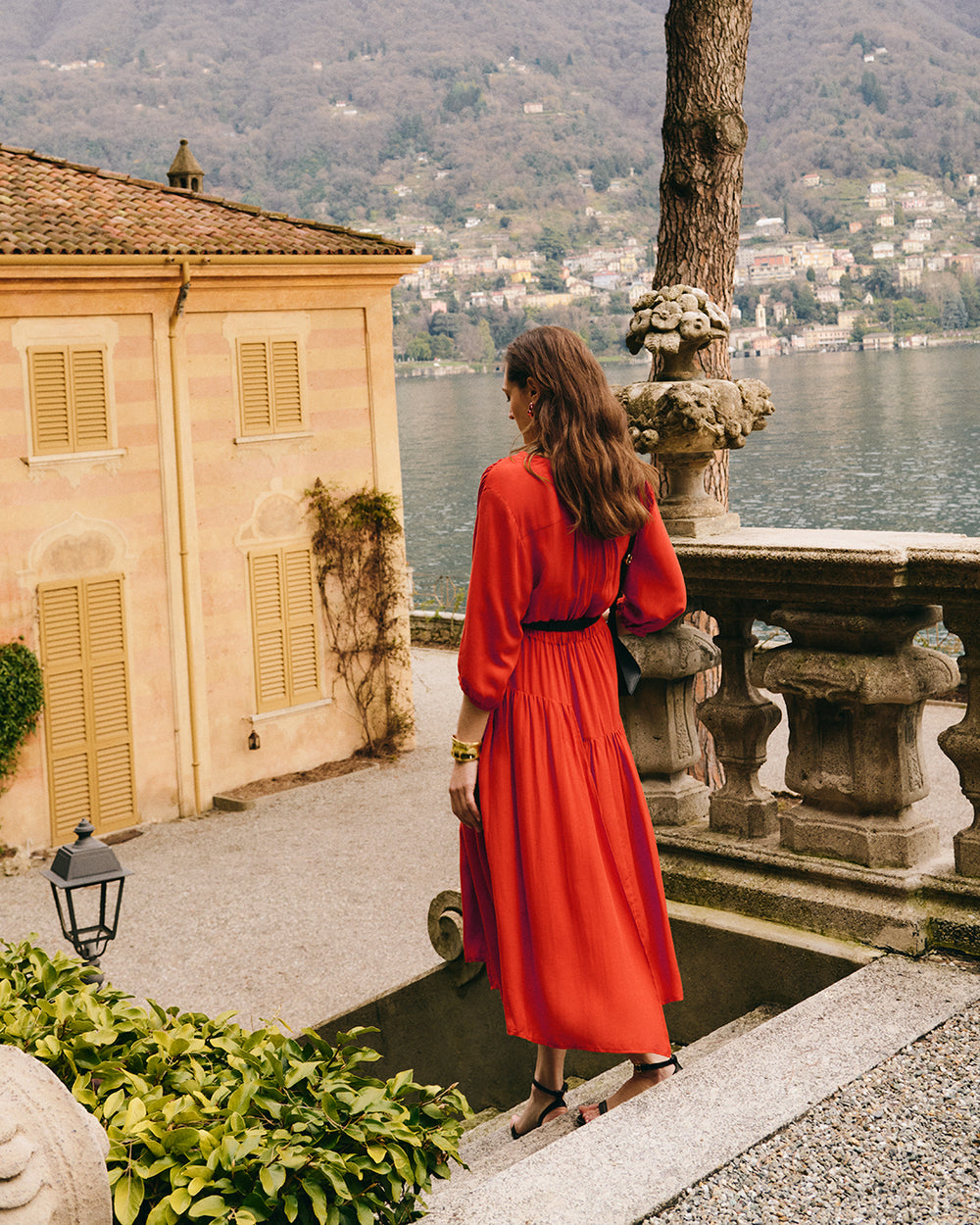 Woman in dress stands near railing overlooking lake and buildings in background.