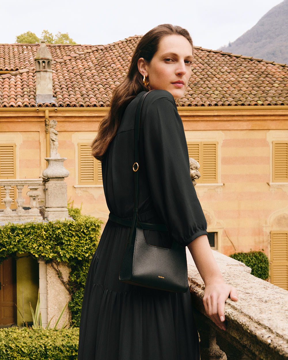 Person in a dress stands by a stone railing with a small bag.