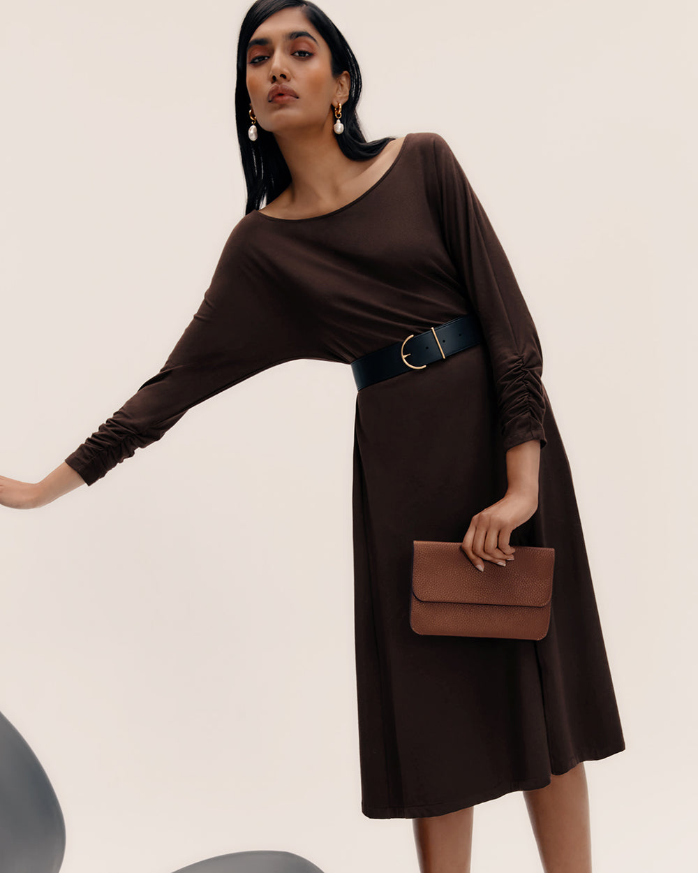 Person in dress with wide belt holding a clutch standing against plain background