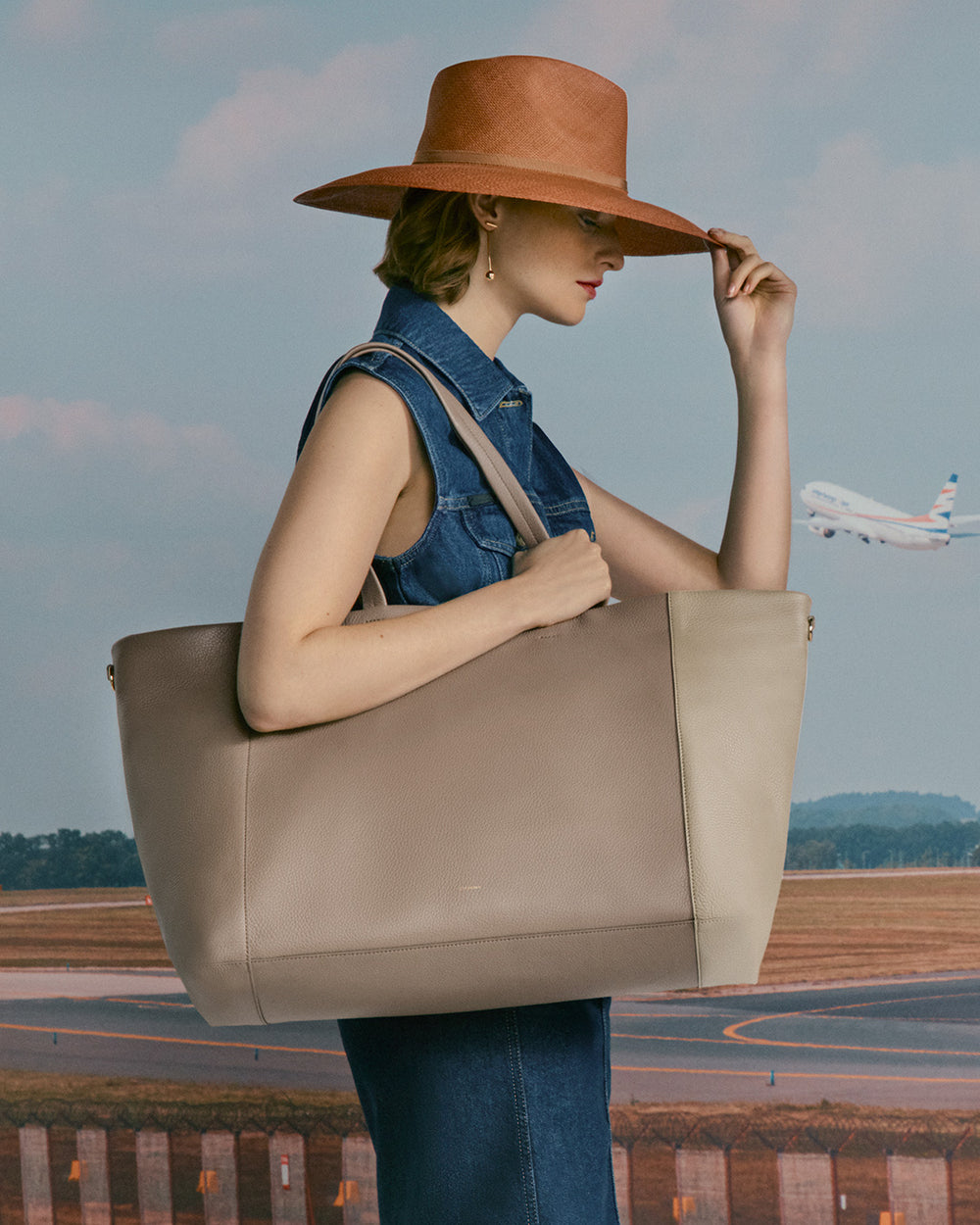 Person holding a large bag standing in front of a runway with a plane in the background, wearing a hat.