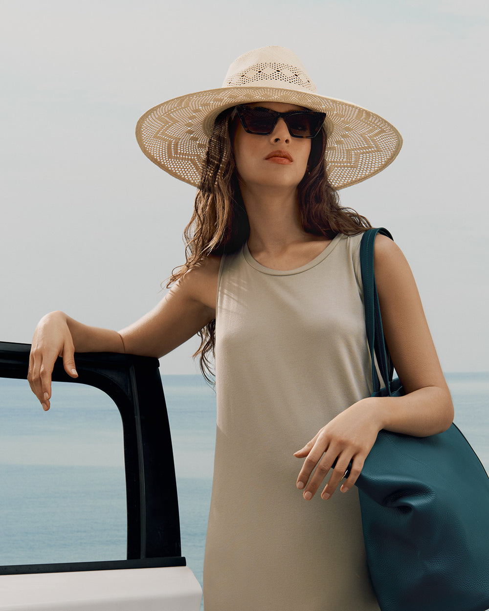 Person with hat and sunglasses standing by a car with a travel bag over their shoulder.