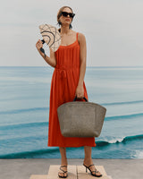 Woman in a dress holding a fan and a bag by the sea.