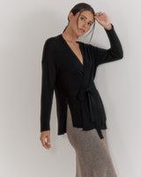 Woman in a belted top and long skirt posing with one hand raised to her head.