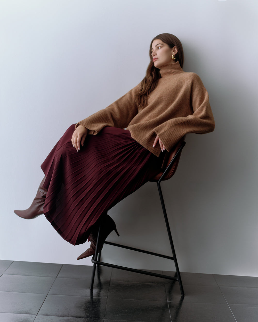Person sitting on a chair wearing a loose sweater and a pleated skirt with boots.