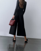 Person holding a purse, wearing wide-leg pants and backless shoes, standing on a tile floor.