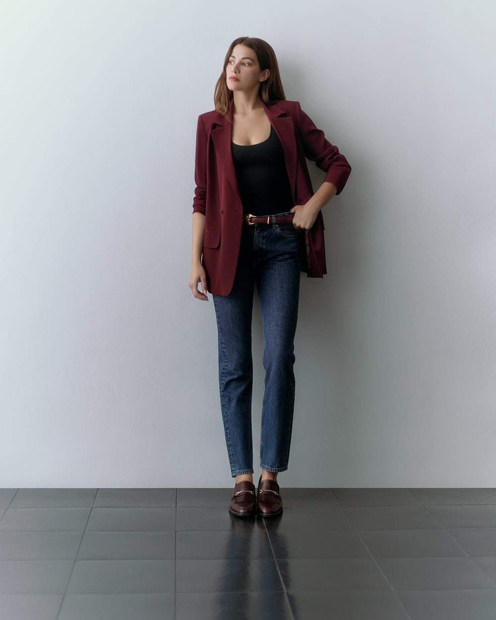 Person stands against a wall, wearing a red blazer, black top, jeans, and loafers.