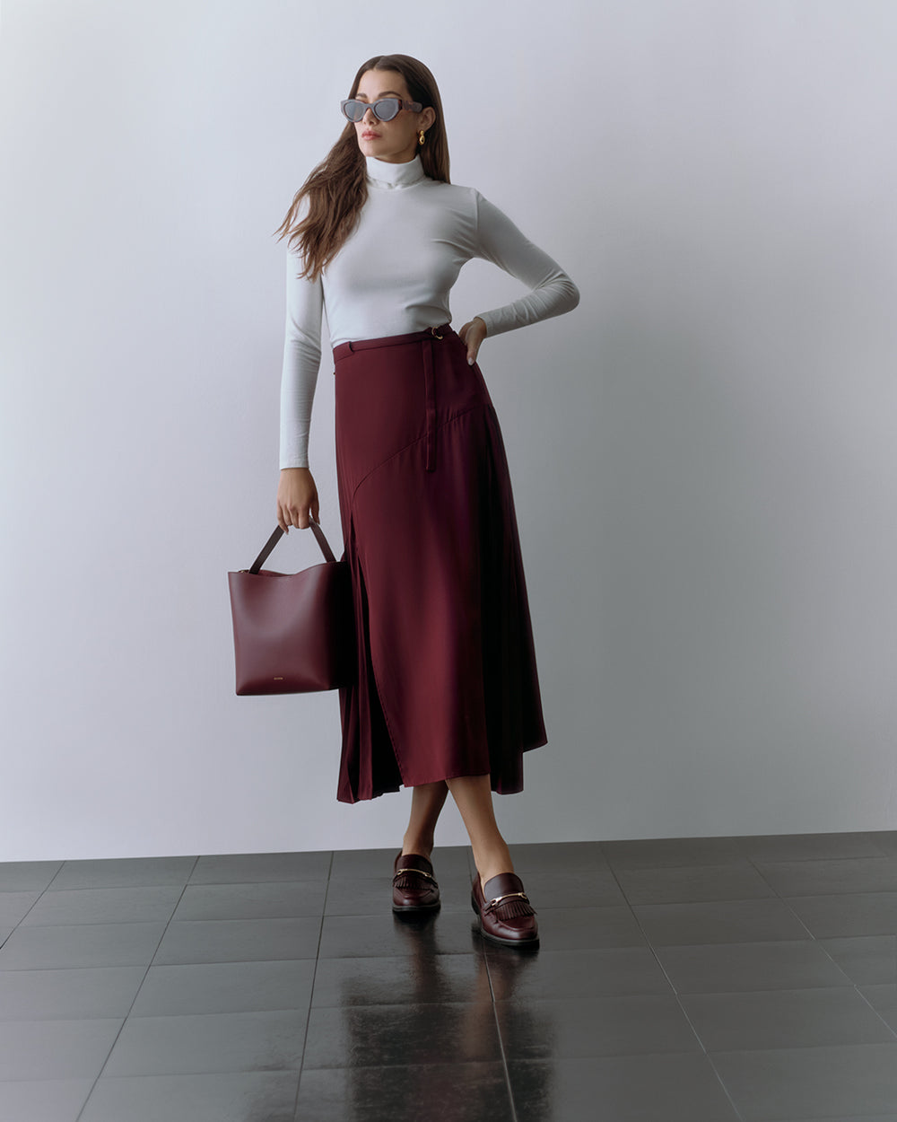 Person wearing a fitted top and long skirt, holding a tote bag and wearing sunglasses.