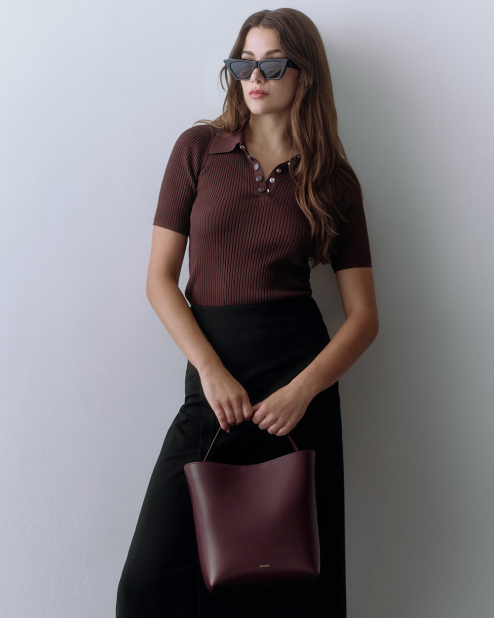 Woman with sunglasses holds a handbag, wearing a short sleeve top and long skirt, standing indoors.
