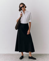 Person wears sunglasses and holds a bag, dressed in a button-up shirt and long skirt.