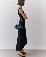 Woman in long dress with shoulder bag and sandals, standing against plain backdrop.