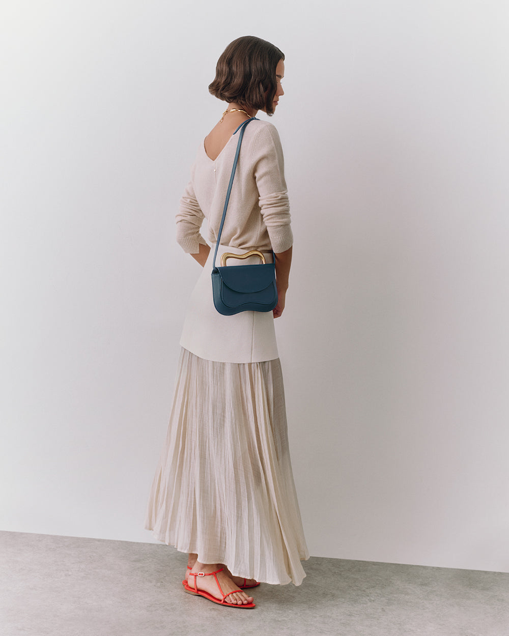 Person standing, wearing long skirt, top, holding small shoulder bag, and wearing sandals.