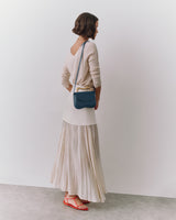 Person standing, wearing long skirt, top, holding small shoulder bag, and wearing sandals.
