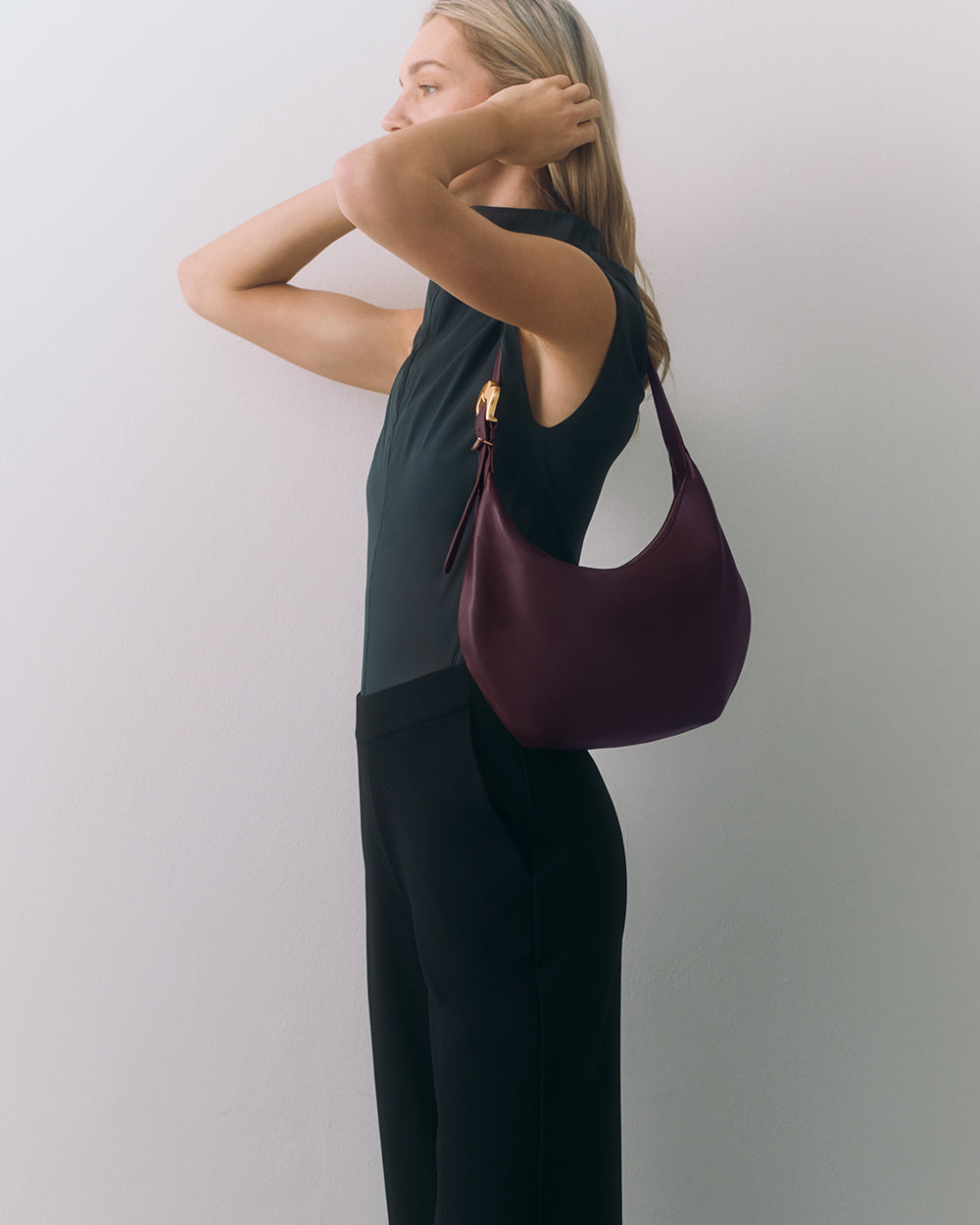Person standing sideways, holding hair, wearing sleeveless top and holding a handbag.