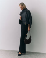 Person in a jacket and pants holding a shoulder bag standing against a plain backdrop.