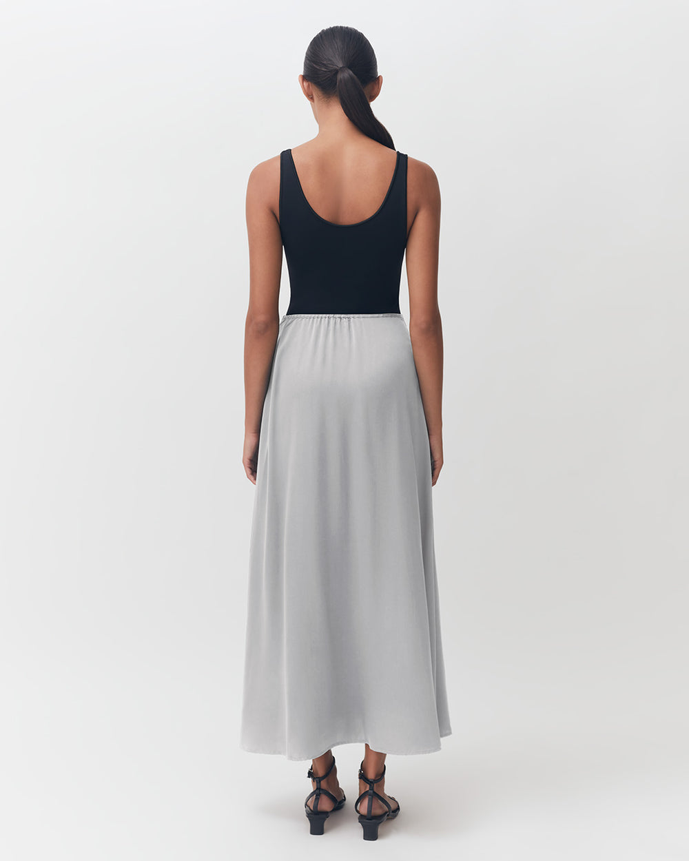 Woman standing facing away, wearing a sleeveless top and long skirt, with her hair tied back.