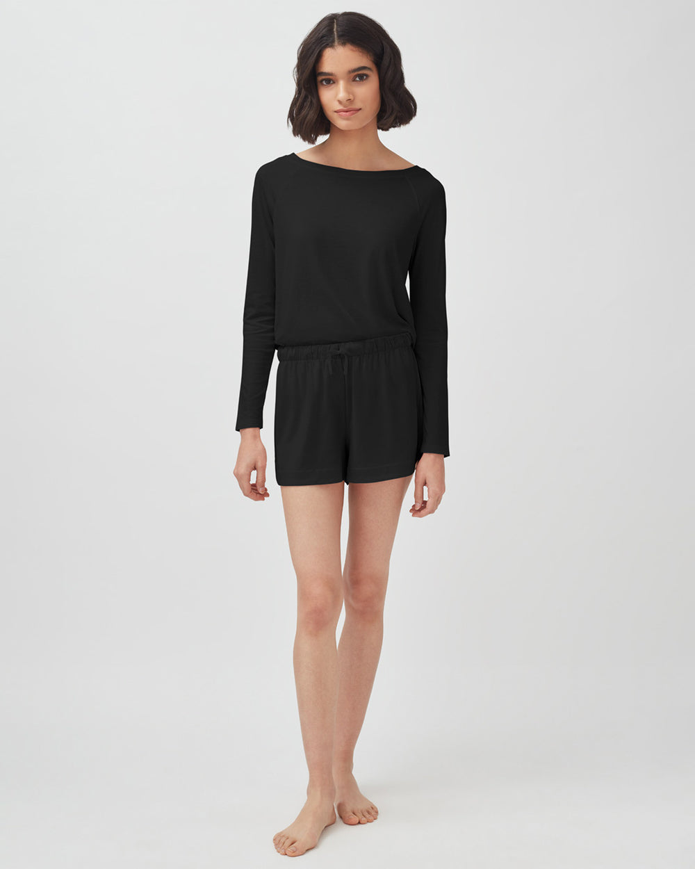 Woman standing in a long-sleeve top and shorts, barefoot.