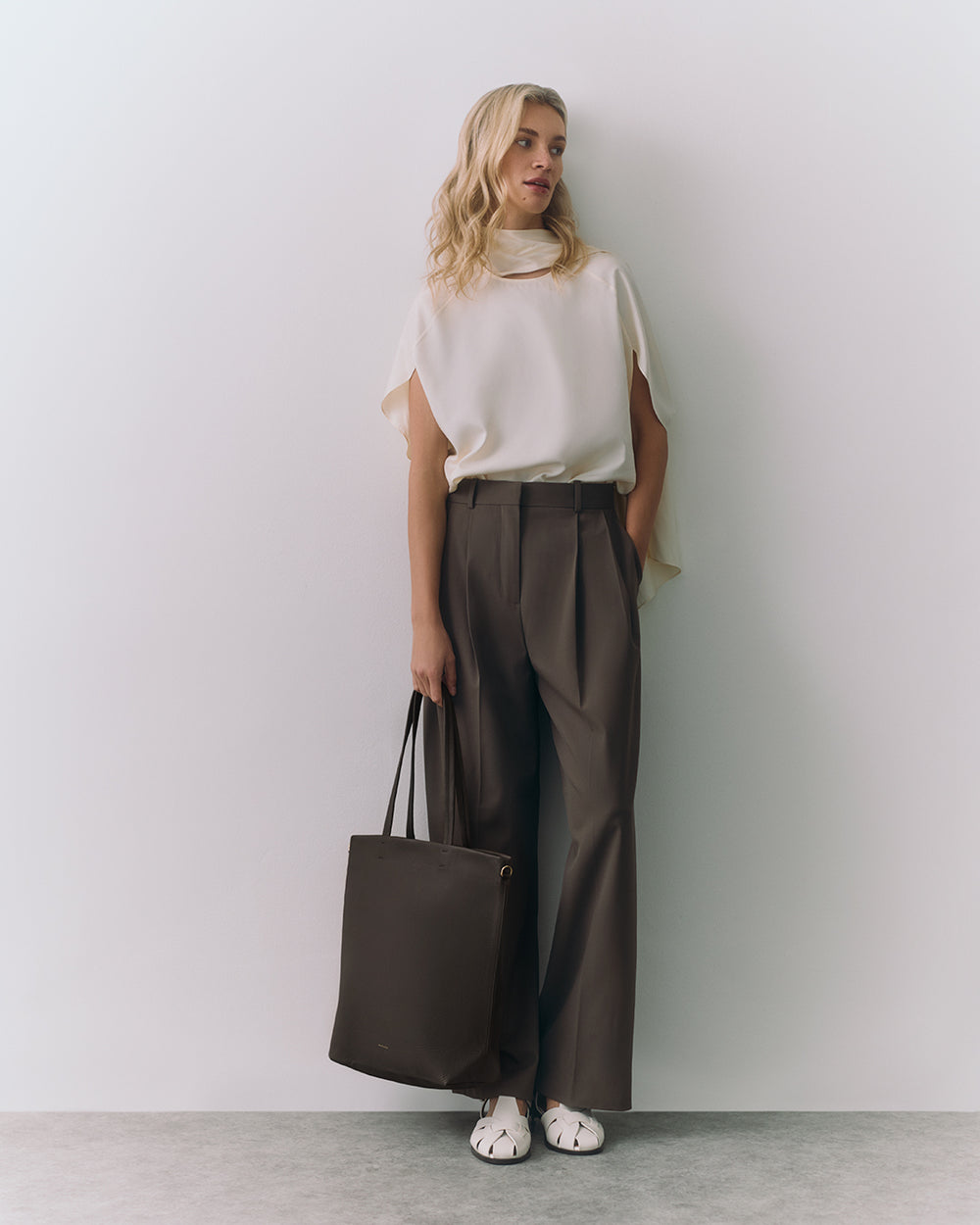 Person standing against a wall holding a tote bag, wearing a top and trousers