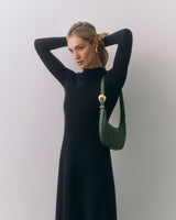 Person in long dress with a shoulder bag adjusts hair, standing against plain background.
