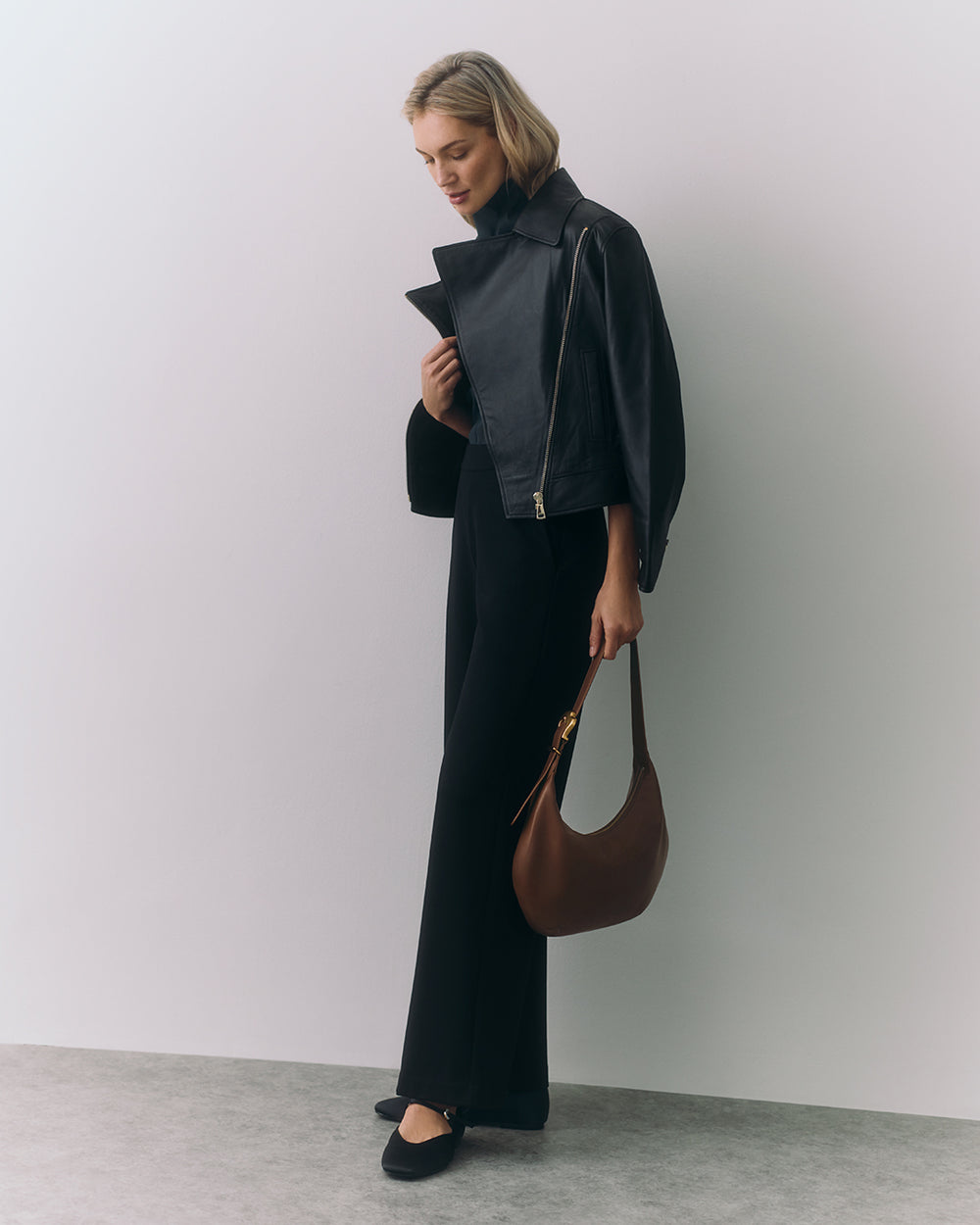 Person in a jacket holding a handbag stands against a plain wall