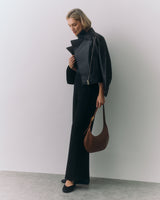 Person in a jacket holding a handbag stands against a plain wall