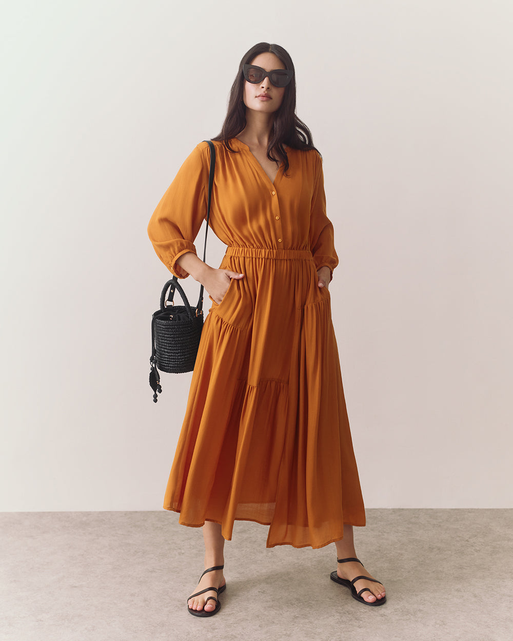 Woman in long dress with sunglasses holding a woven bag, standing indoors.