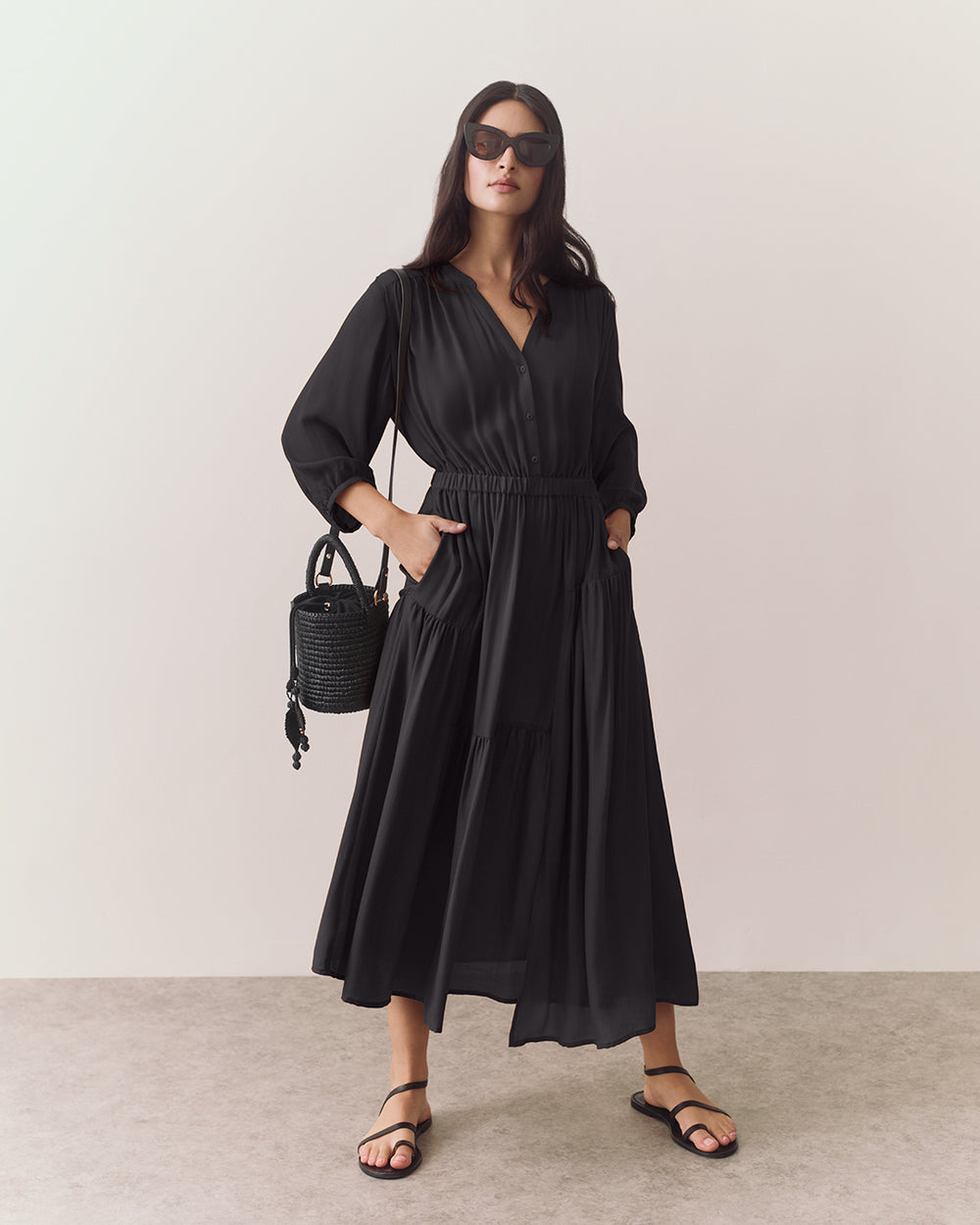 Woman in long dress with sunglasses holds a woven handbag and stands on a plain background.
