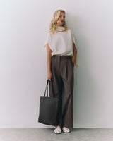 Person stands against wall holding tote bag, wearing loose top and trousers.