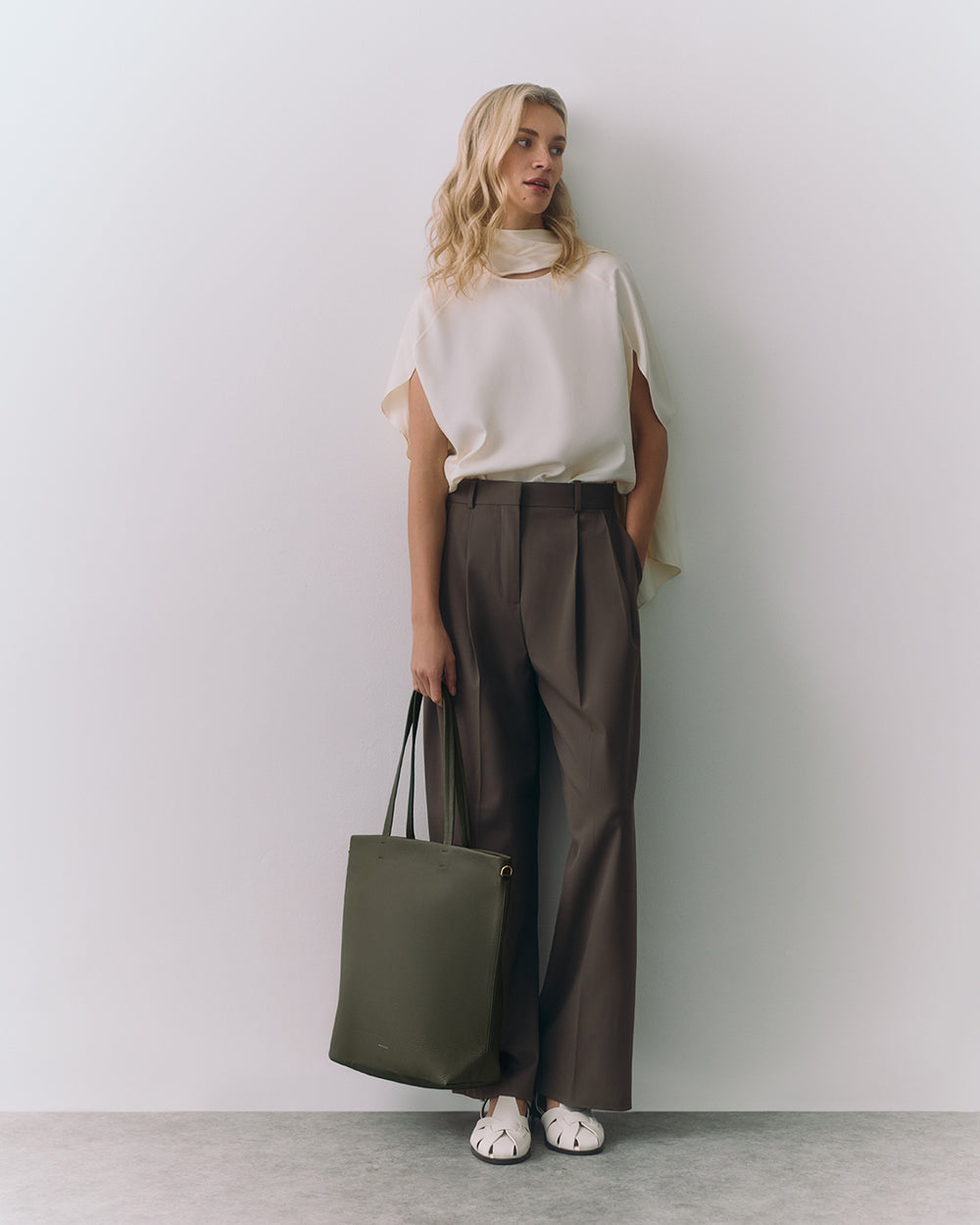 Woman standing against wall holding a tote bag and wearing a sleeveless top with wide-leg pants.