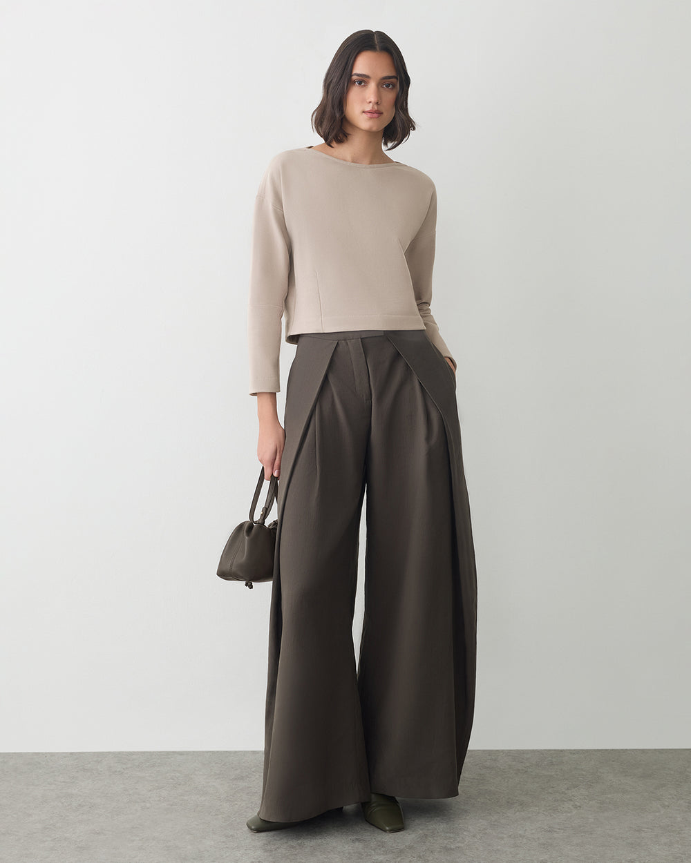 Person wearing a long-sleeve top and wide-leg pants holding a small bag in a studio setting.