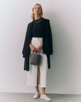 Person wearing a top, cape, pants, holding a small bag, standing against a plain backdrop.