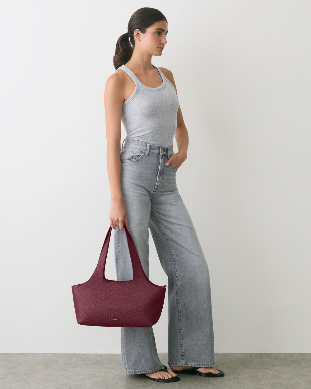 Person in tank top and jeans holding a handbag in a minimal setting.