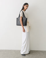 Woman in long dress with croc-embossed leather tote stands against a plain wall.