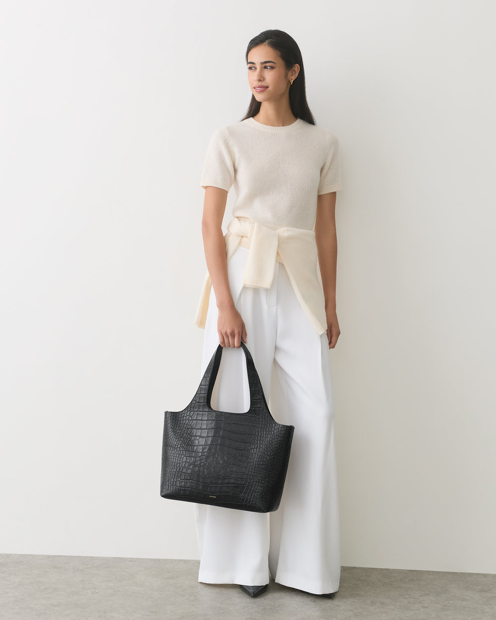 Model holding croc-embossed leather tote bag, wearing white pants and shirt.
