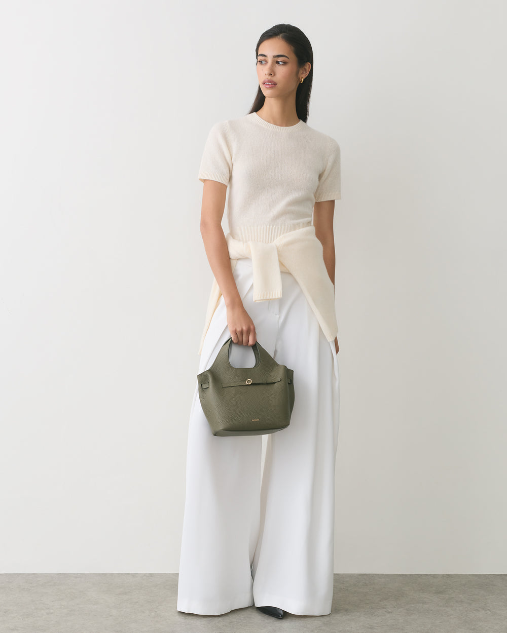 Woman in wide-leg pants holds small handbag standing against plain backdrop.