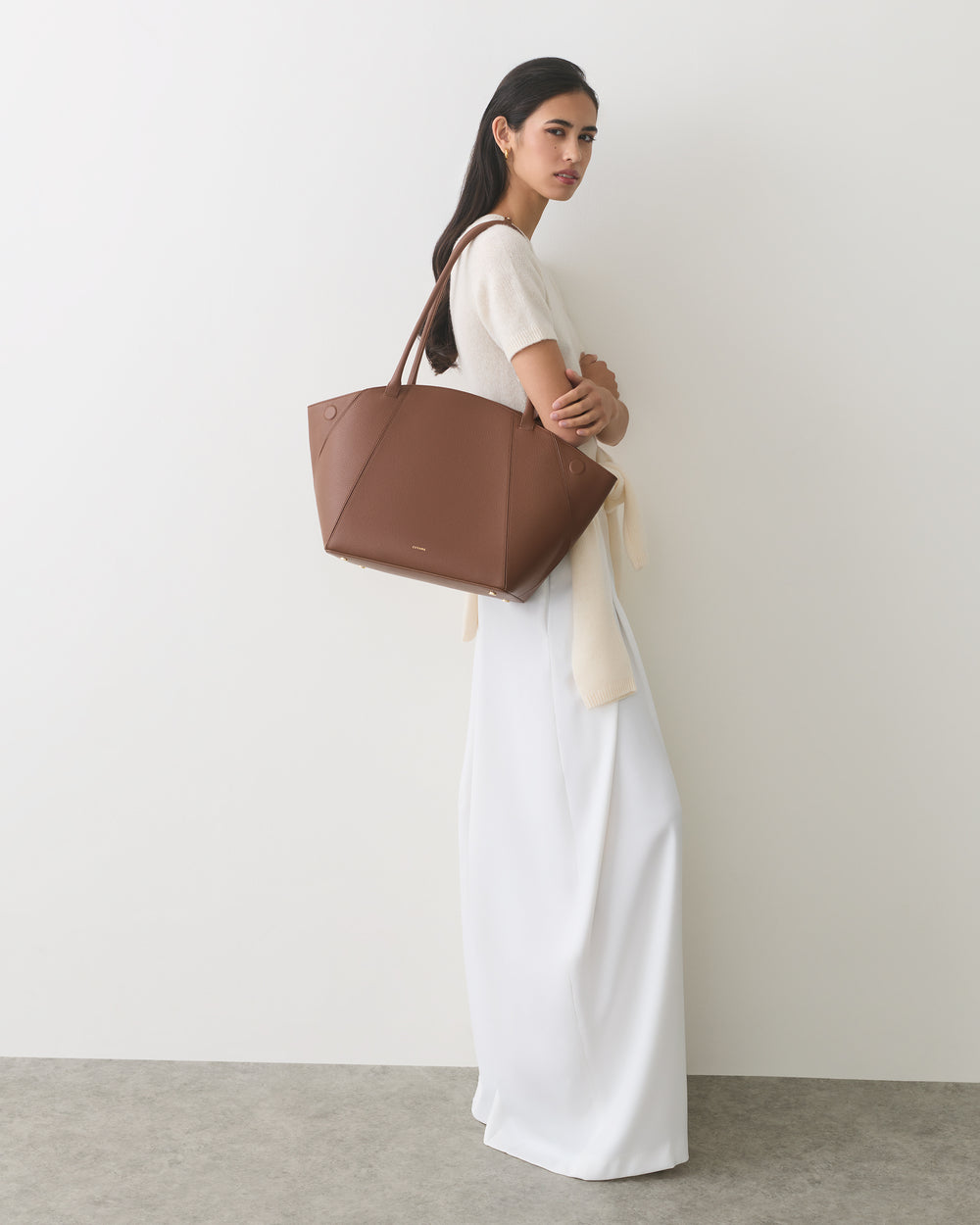 Person standing with large tote bag, wearing a long dress, against a plain background.