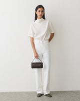 Person standing against plain wall holding a small handbag wearing a short-sleeve top and pants.
