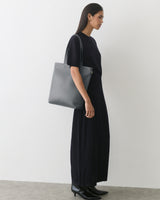 Person in dress holding tote bag, standing against plain wall.