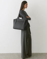 Person in long dress holding large shoulder bag standing against plain background.