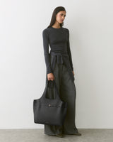 Person with long sleeves, wide pants, holding a large handbag, standing against plain backdrop.