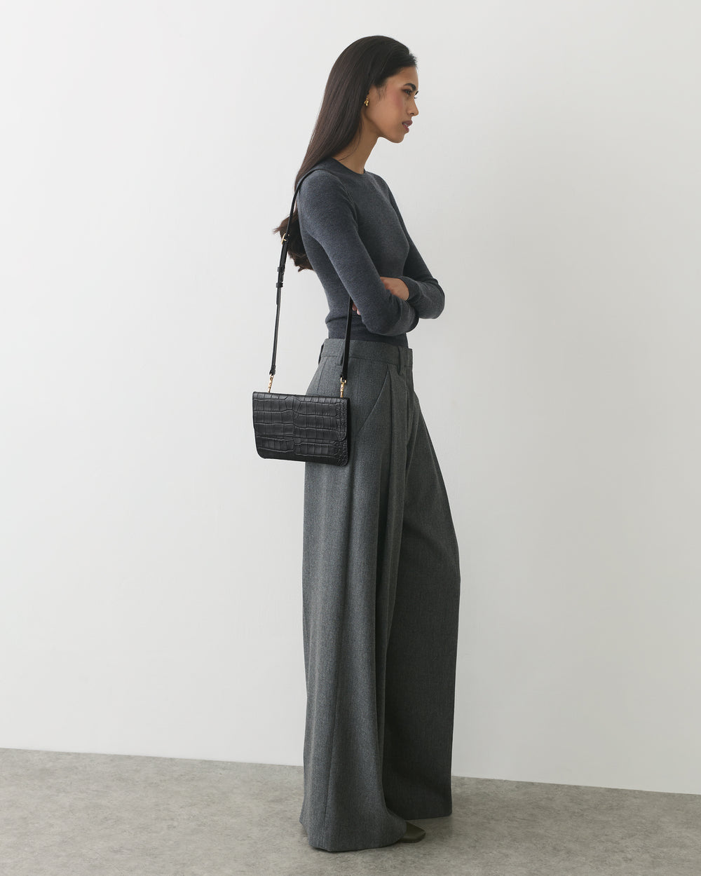 Person standing with arms crossed, wearing wide-leg trousers and a croc-embossed leather bag.