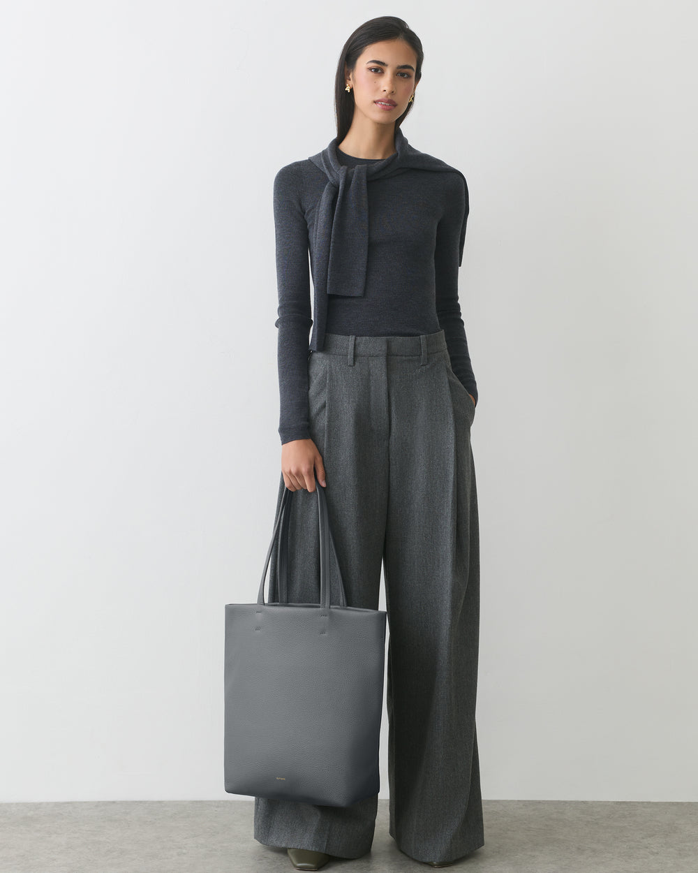 Person standing, wearing long-sleeve top and wide pants, holding a tote bag.