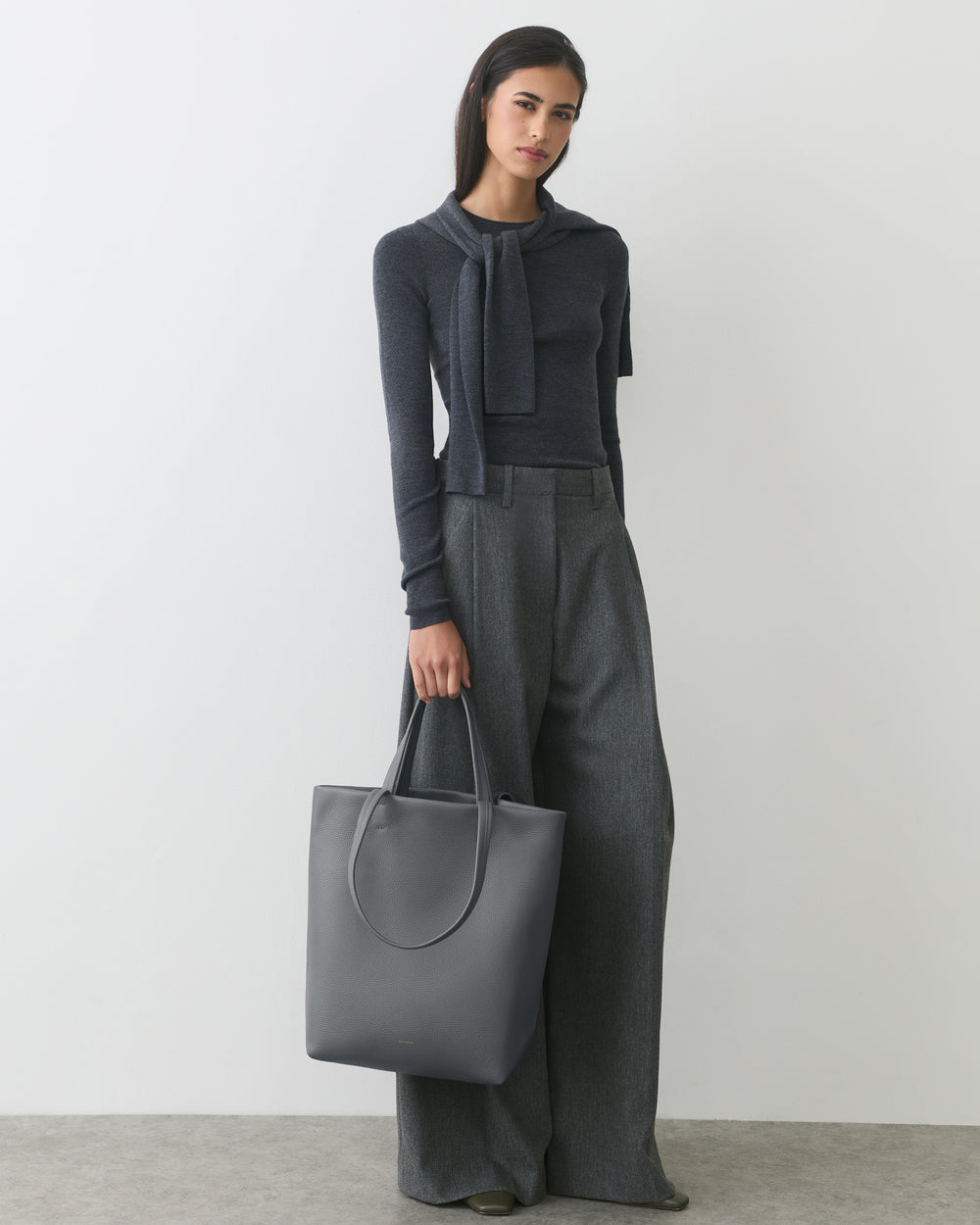 Person standing with a large tote bag, wearing a tied sweater and wide-leg pants.