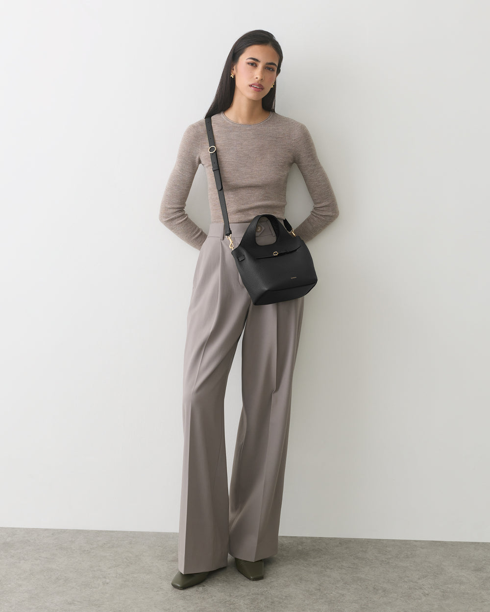 Person standing with a handbag, wearing a sweater and wide-leg pants.
