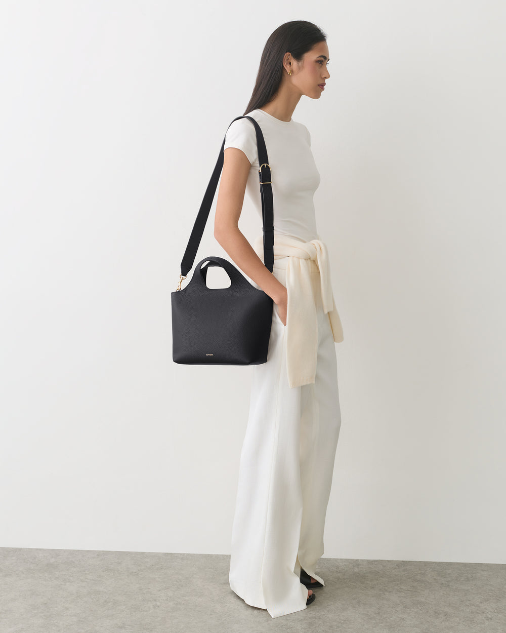 Person standing, wearing a long dress, holding a handbag and sweater tied around waist.