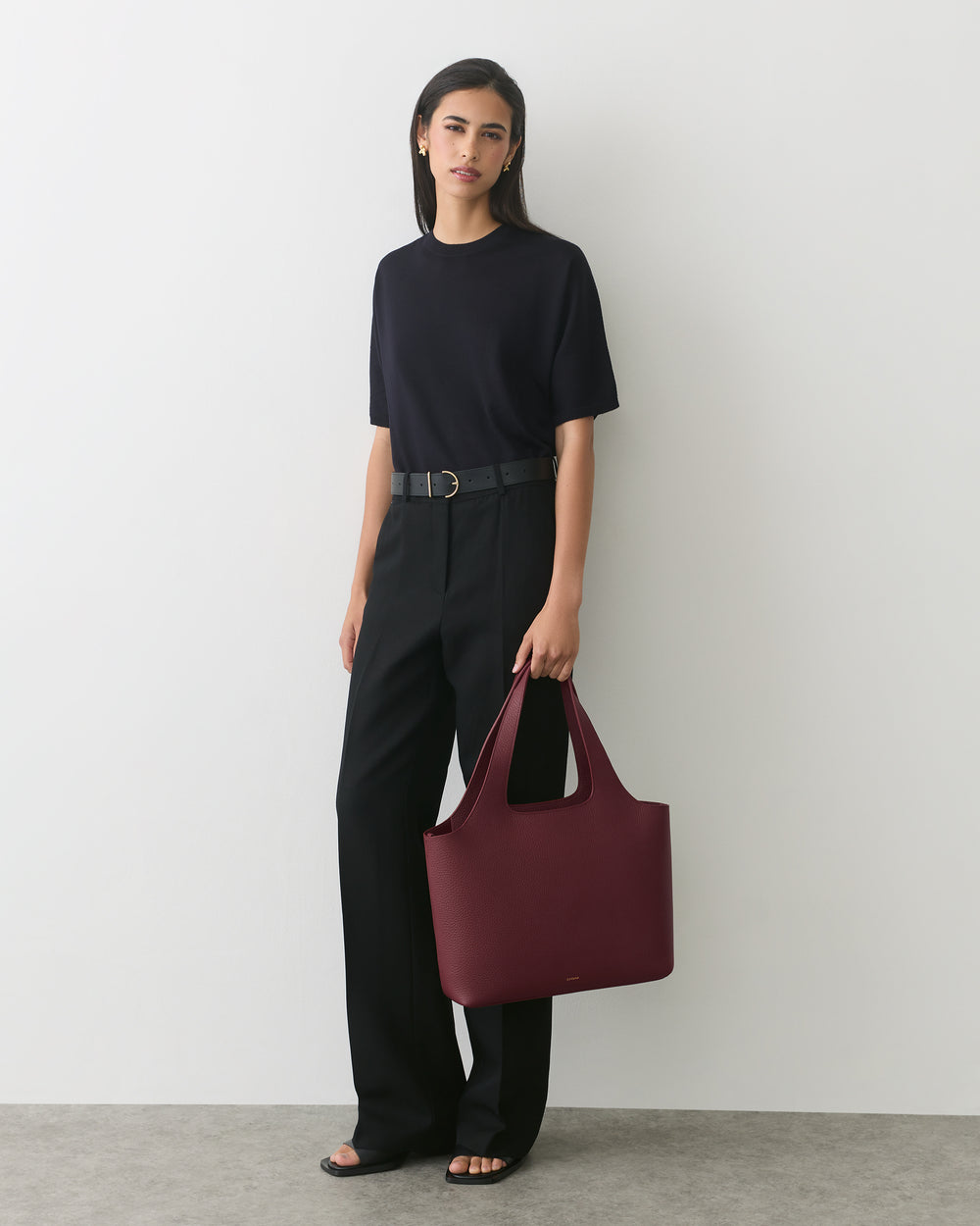 Person in a T-shirt and pants holding a large handbag standing against a plain background.