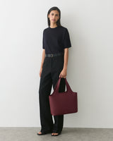 Person in a T-shirt and pants holding a large handbag standing against a plain background.