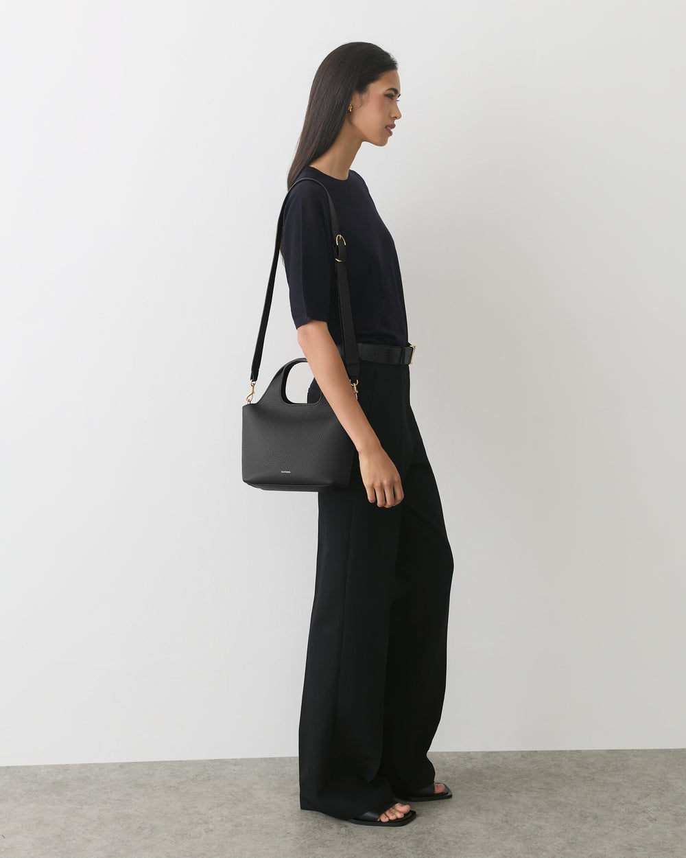 Model wearing a top and pants, holding a bag with a long strap.